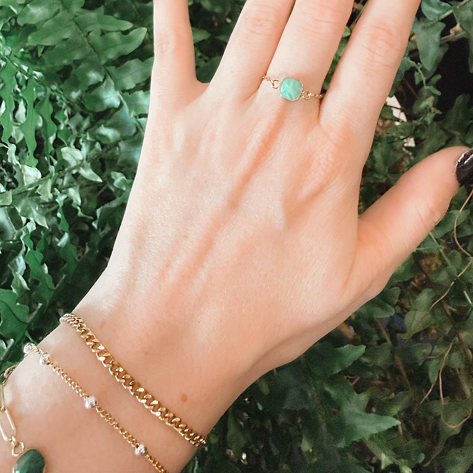 A close-up of a hand with dark manicured nails wearing a delicate gold ring with a round light green stone, set against a background of green foliage at Azelea Society.