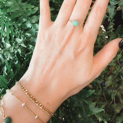 A close-up of a hand with dark manicured nails wearing a delicate gold ring with a round light green stone, set against a background of green foliage at Azelea Society.