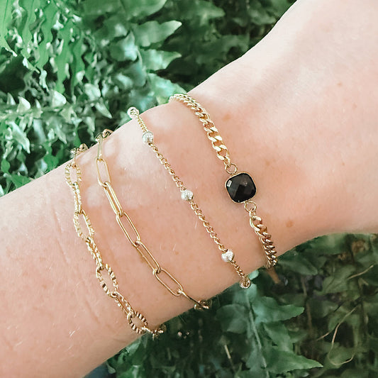A close-up of a person's wrist wearing three delicate gold bracelets, including a chain bracelet with a small black square connector, set against a background of green foliage from Azelea Society