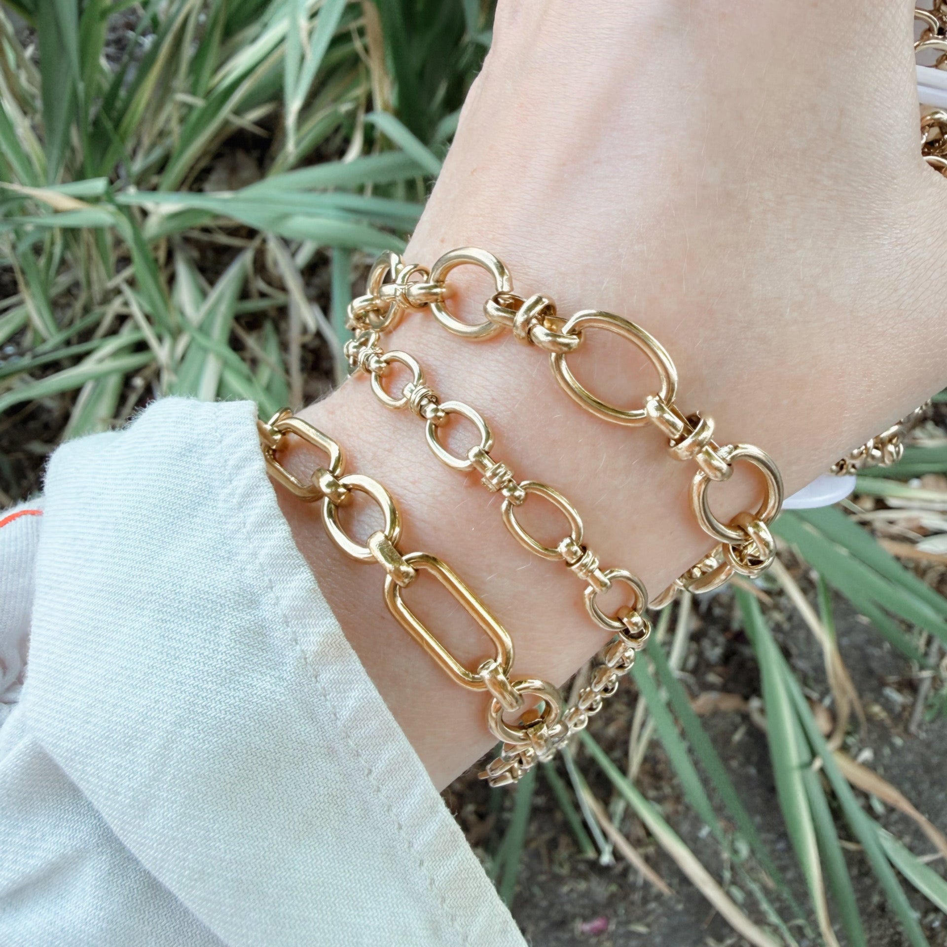 A close-up of a person's wrist at Azelea Society wearing three layered gold chain bracelets with mixed link shapes, set against a background of green plants and a white sleeve.