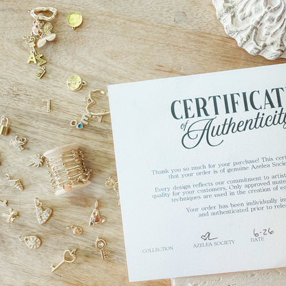 A flat-lay photograph of a white 'Certificate of Authenticity' card from 'Azelea Society' on a light wood background, surrounded by various gold jewelry charms and a gold ring.